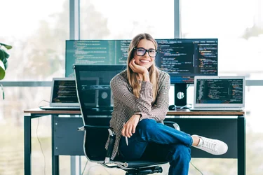 Lächelnde Softwareentwicklerin sitzt vor mehreren Monitoren mit Programmcode.