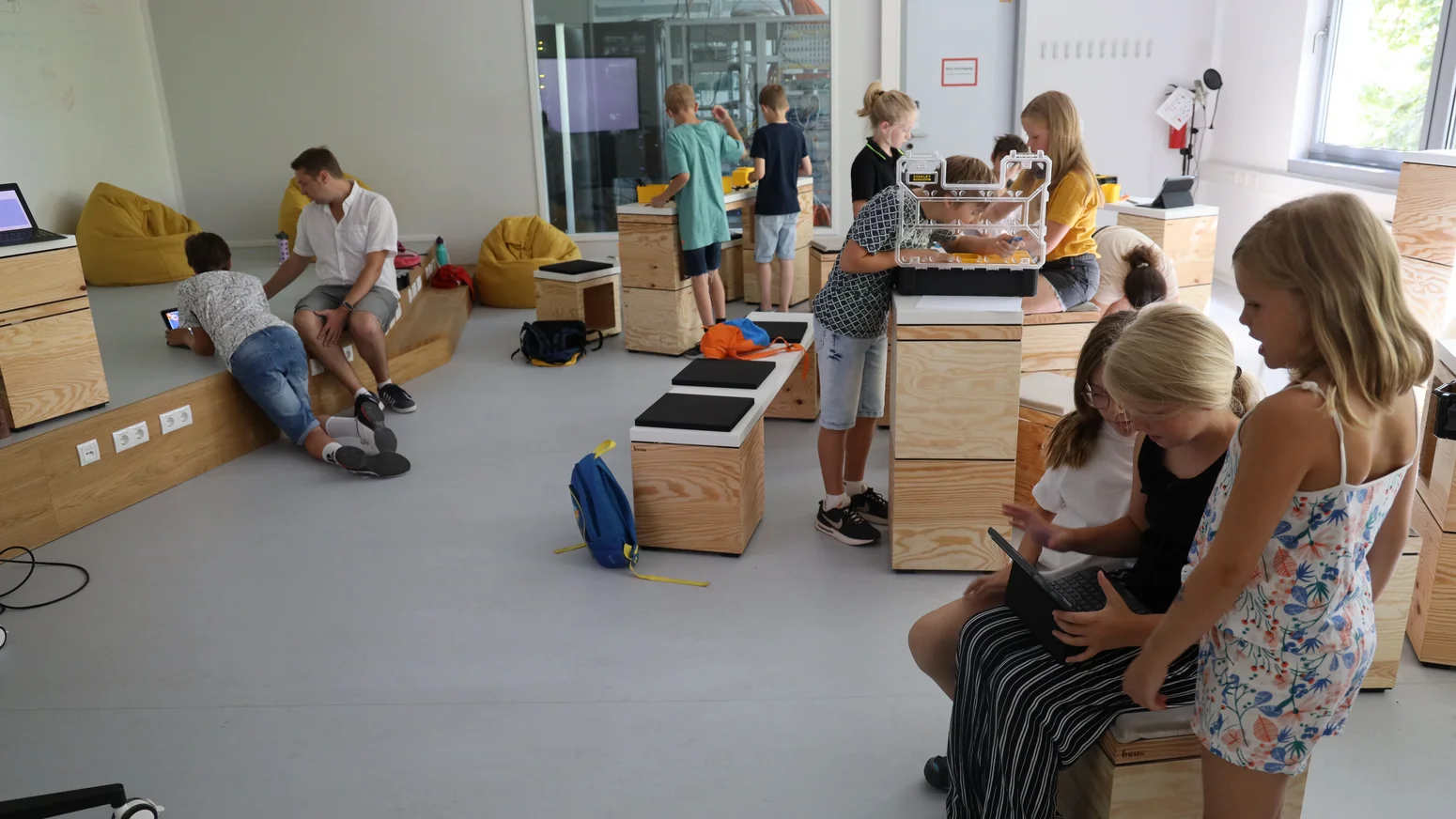 Kinder arbeiten in einem hellen Lernraum mit Holzmöbeln an Laptops und Tablets, im Hintergrund ein Serverschrank hinter Glas, ein Betreuer sitzt auf einer Sitzbank.